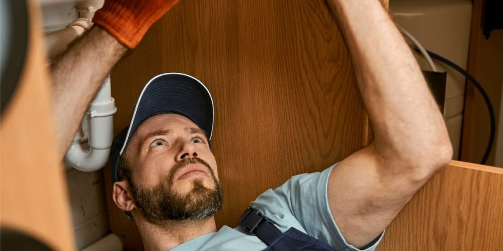 male-plumber-fixing-sink-pipe-in-kitchen-VNBHAW6